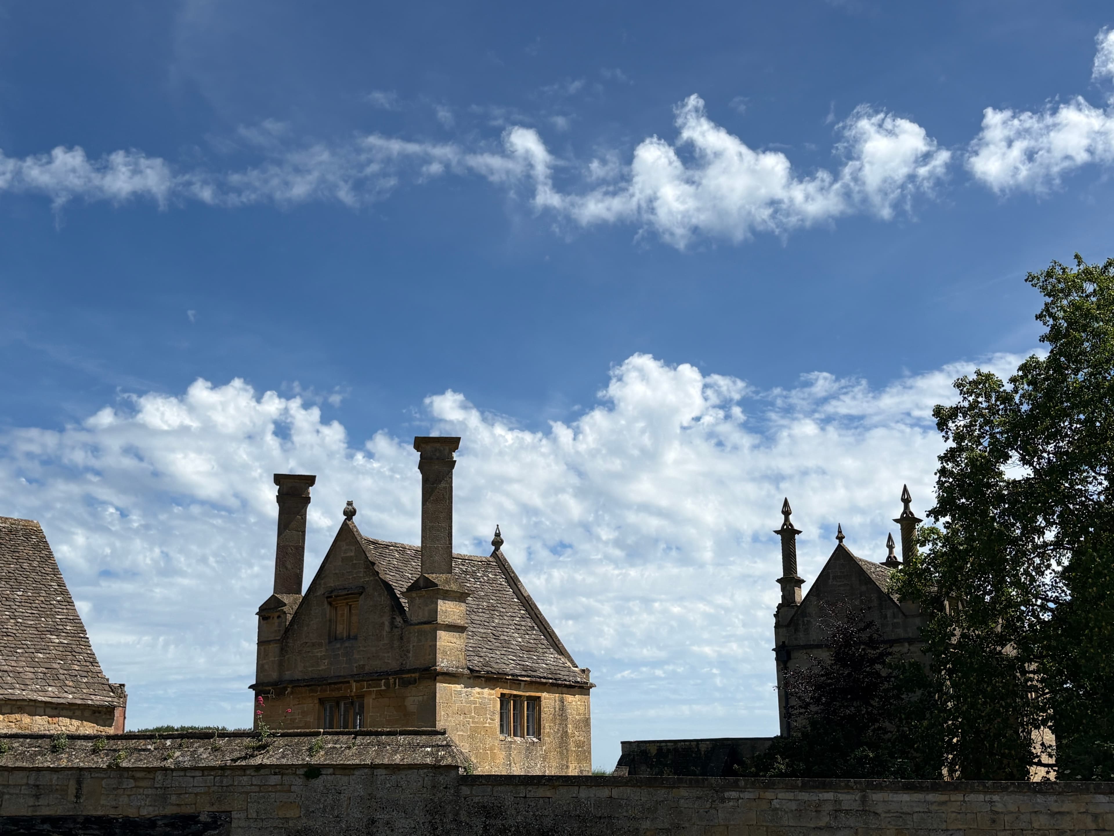 Cotswold-stone gables and clustered chimneys of the Court Barn buildings rising above the boundary wall against a summer sky