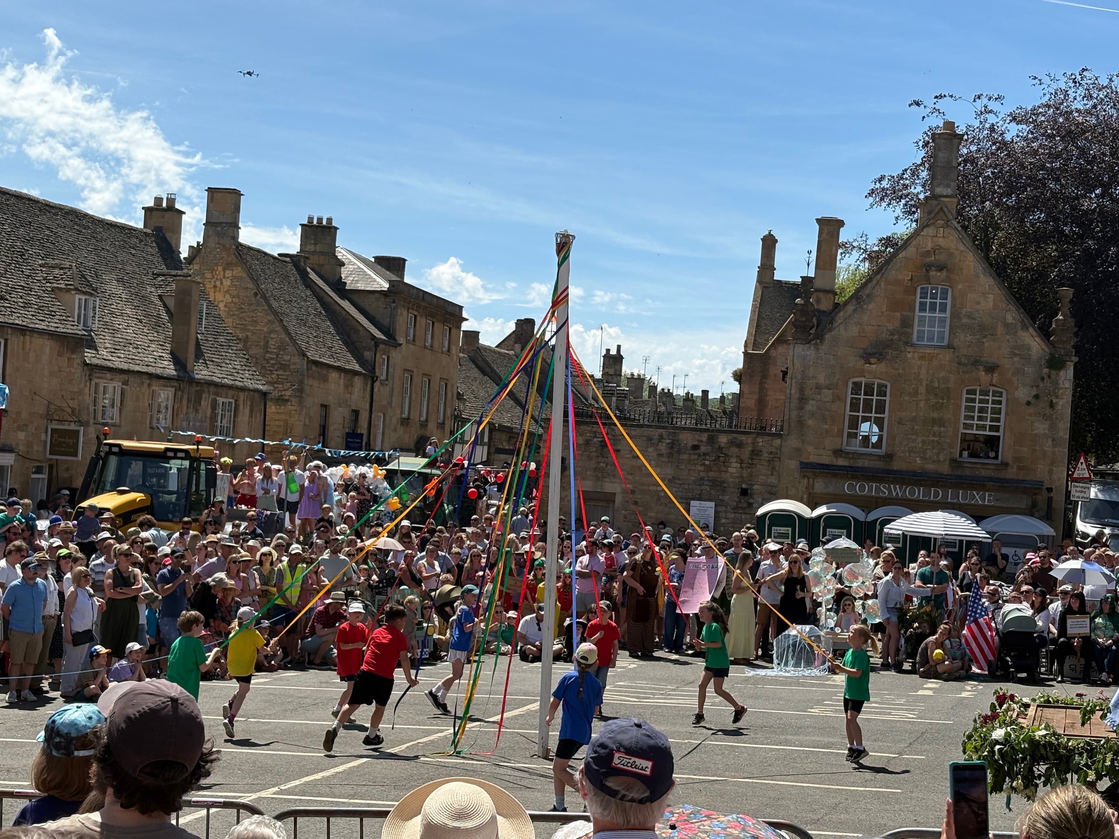 Children dancing around the maypole at the Scuttlebrook Wake in Chipping Campden's Market Square, with the Cotswold-stone high street and a Cotswold Life van behind