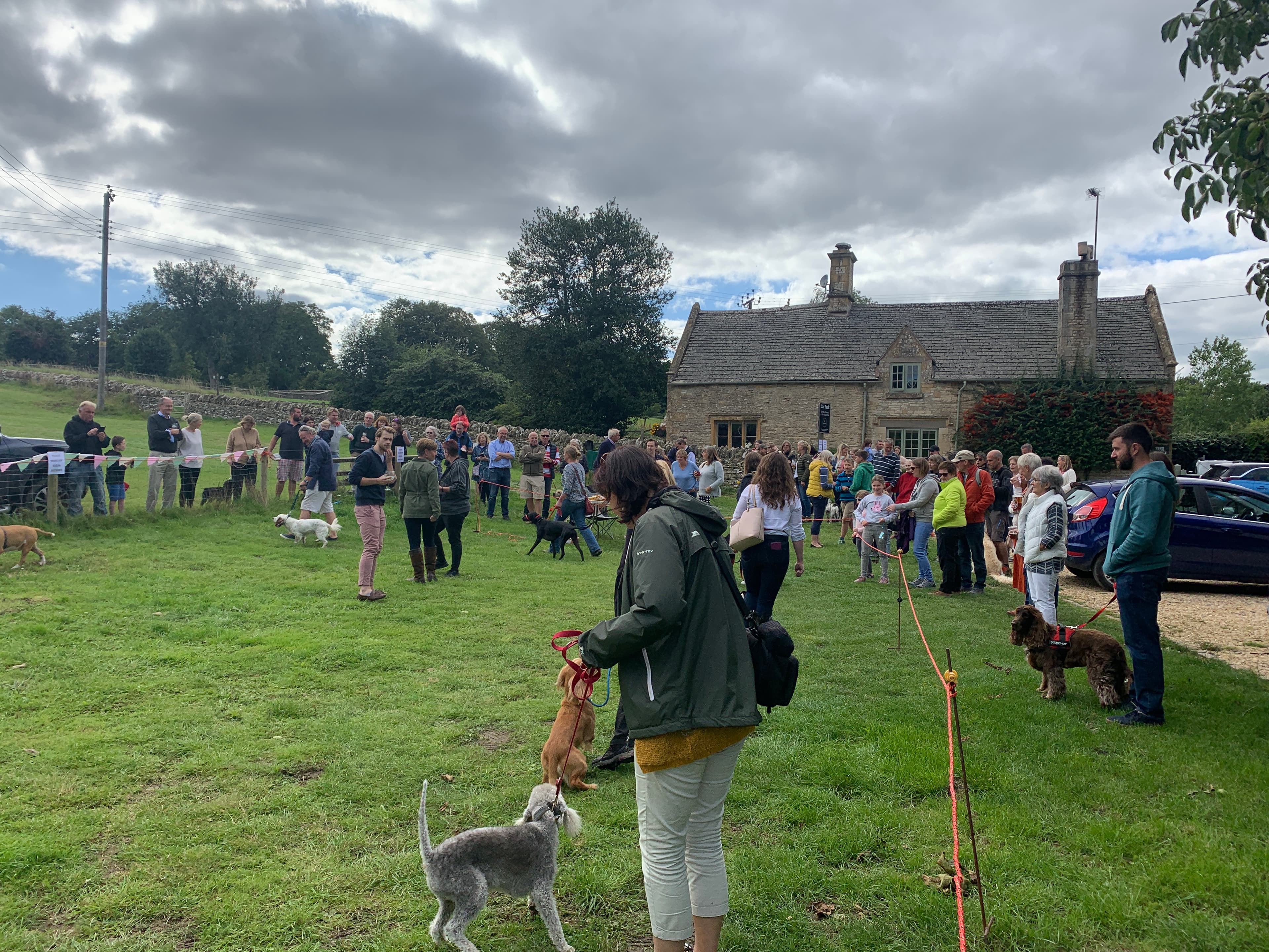 Villagers and dogs gathered on the green at a Notgrove community event, with a Cotswold-stone cottage behind