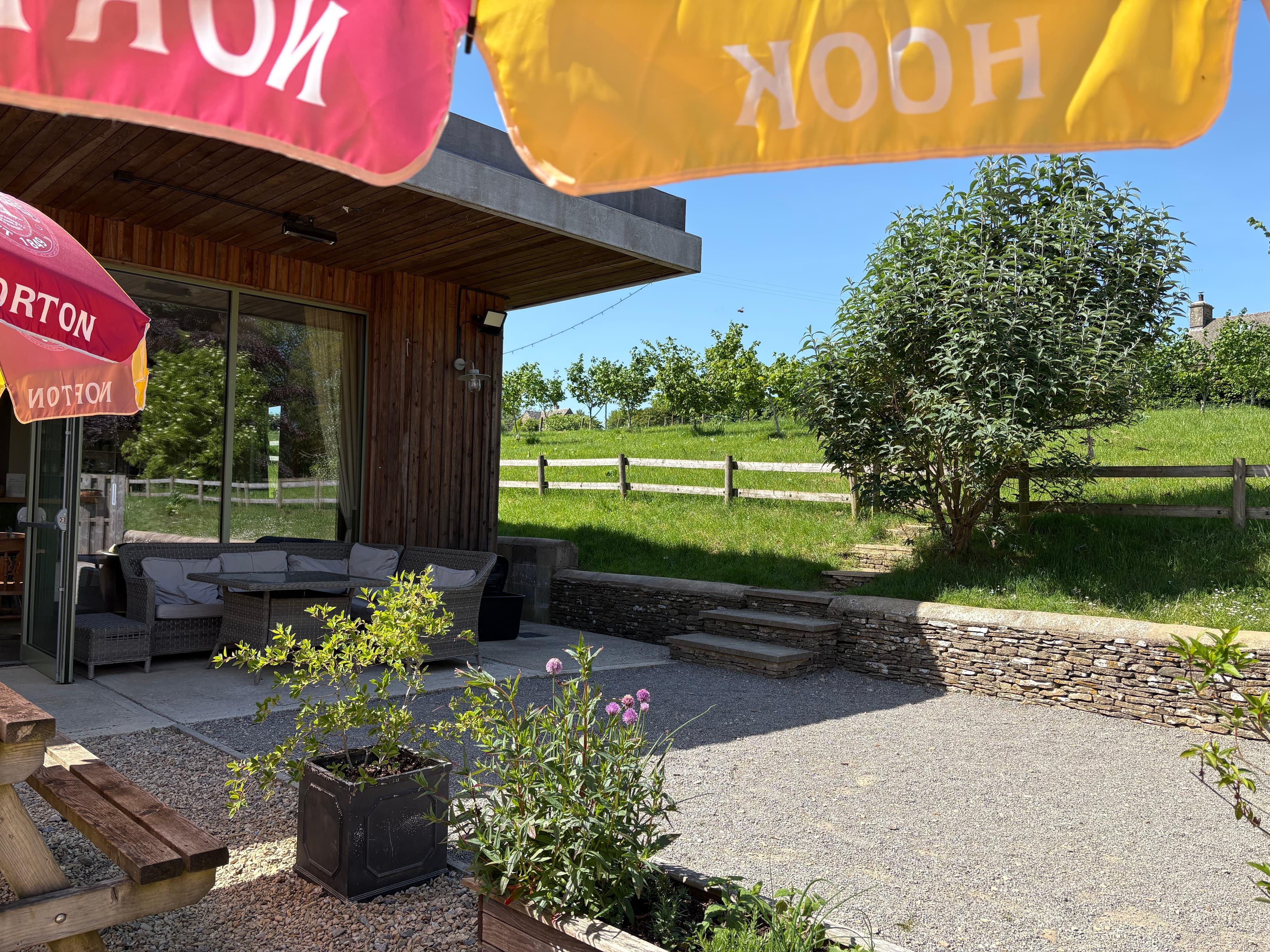 Outdoor seating area on the Notgrove Estate under NOTGROVE pennants, with picnic tables and an orchard beyond