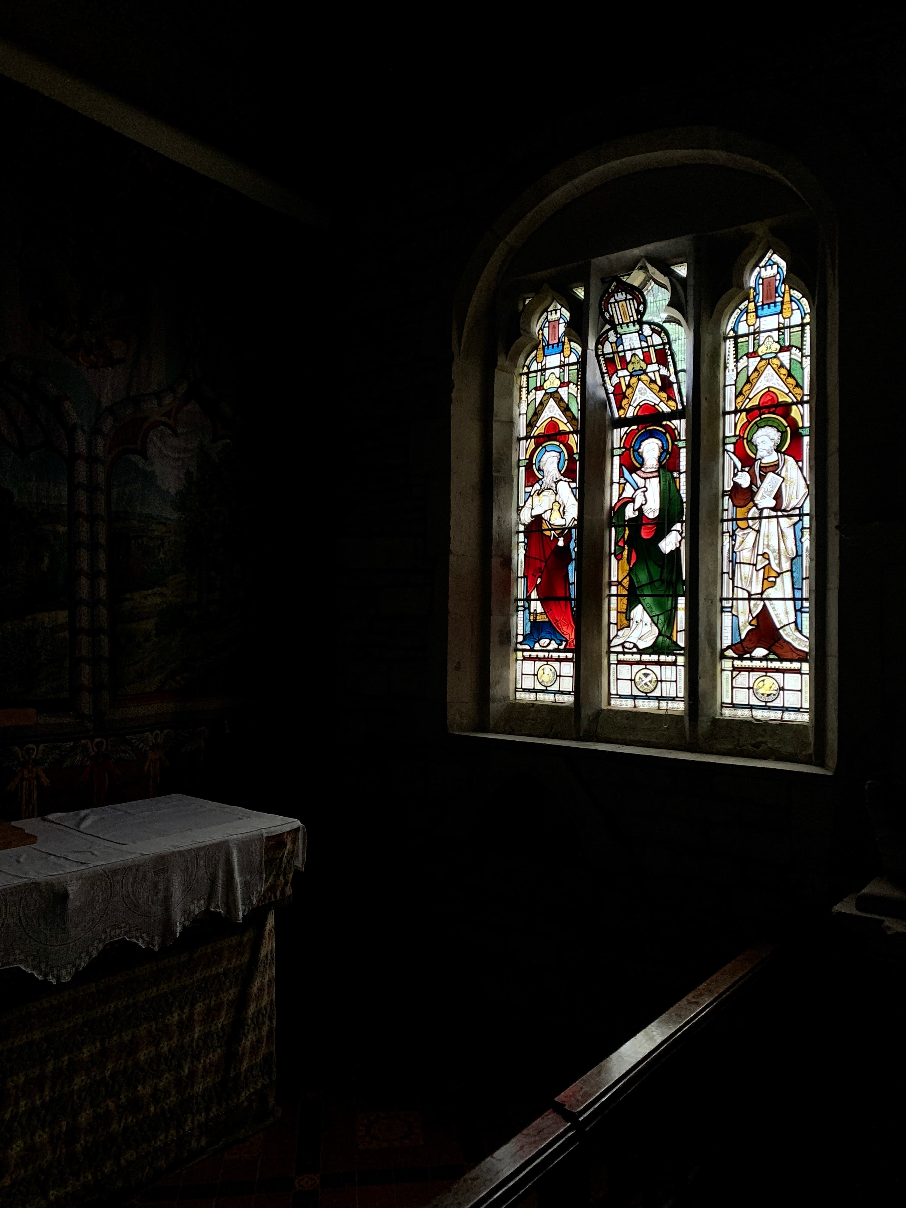 A three-light stained glass window of haloed saints catching the light in the otherwise dark interior of St Bartholomew's, Notgrove