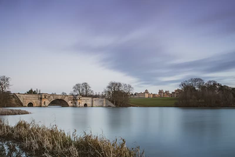 Blenheim Palace's Grand Bridge across the lake at dusk, with the palace silhouette in the distance