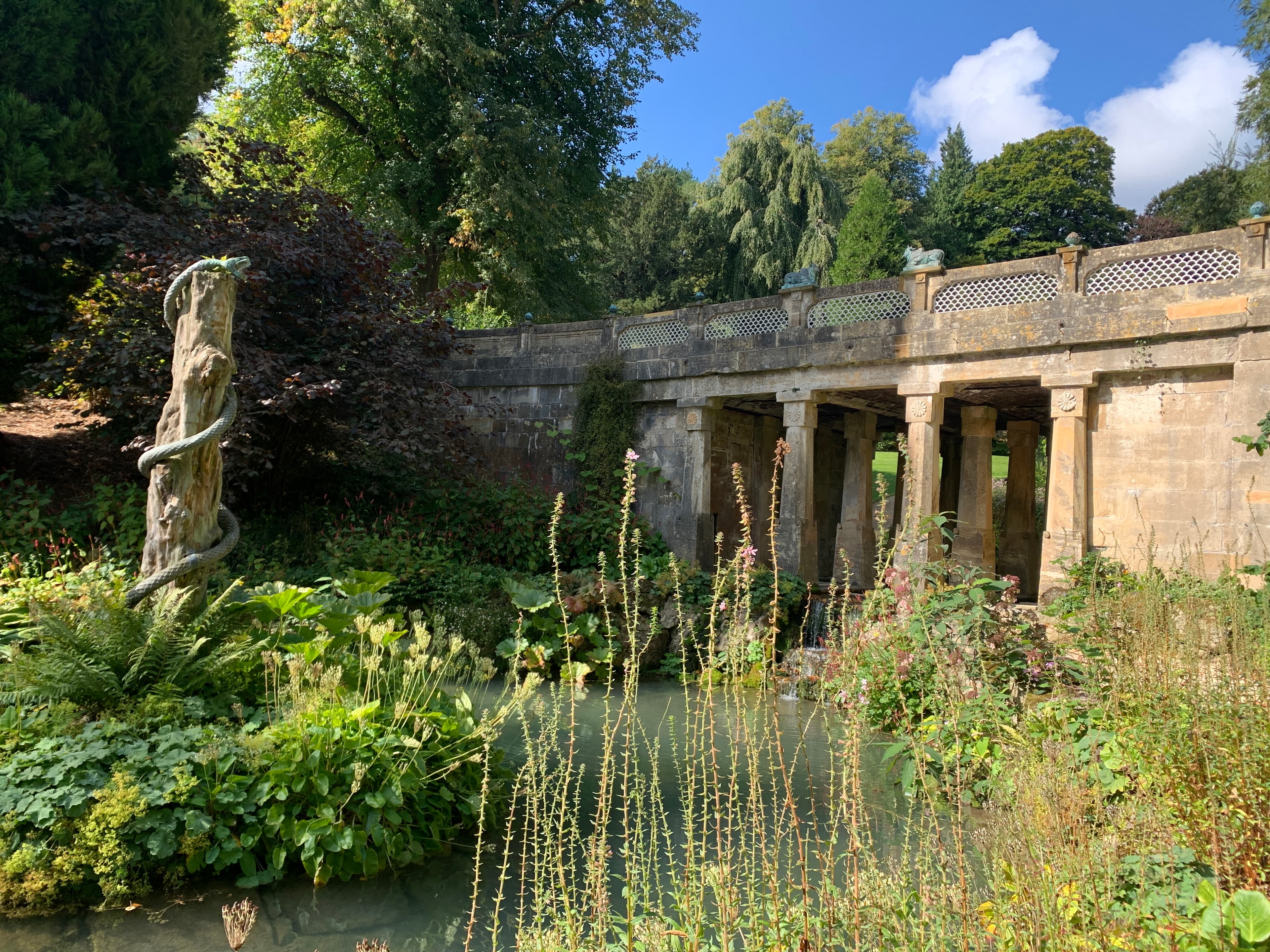 A stone bridge over Sezincote's water garden, with the serpent-entwined pillar on the left and lush planting around the pool