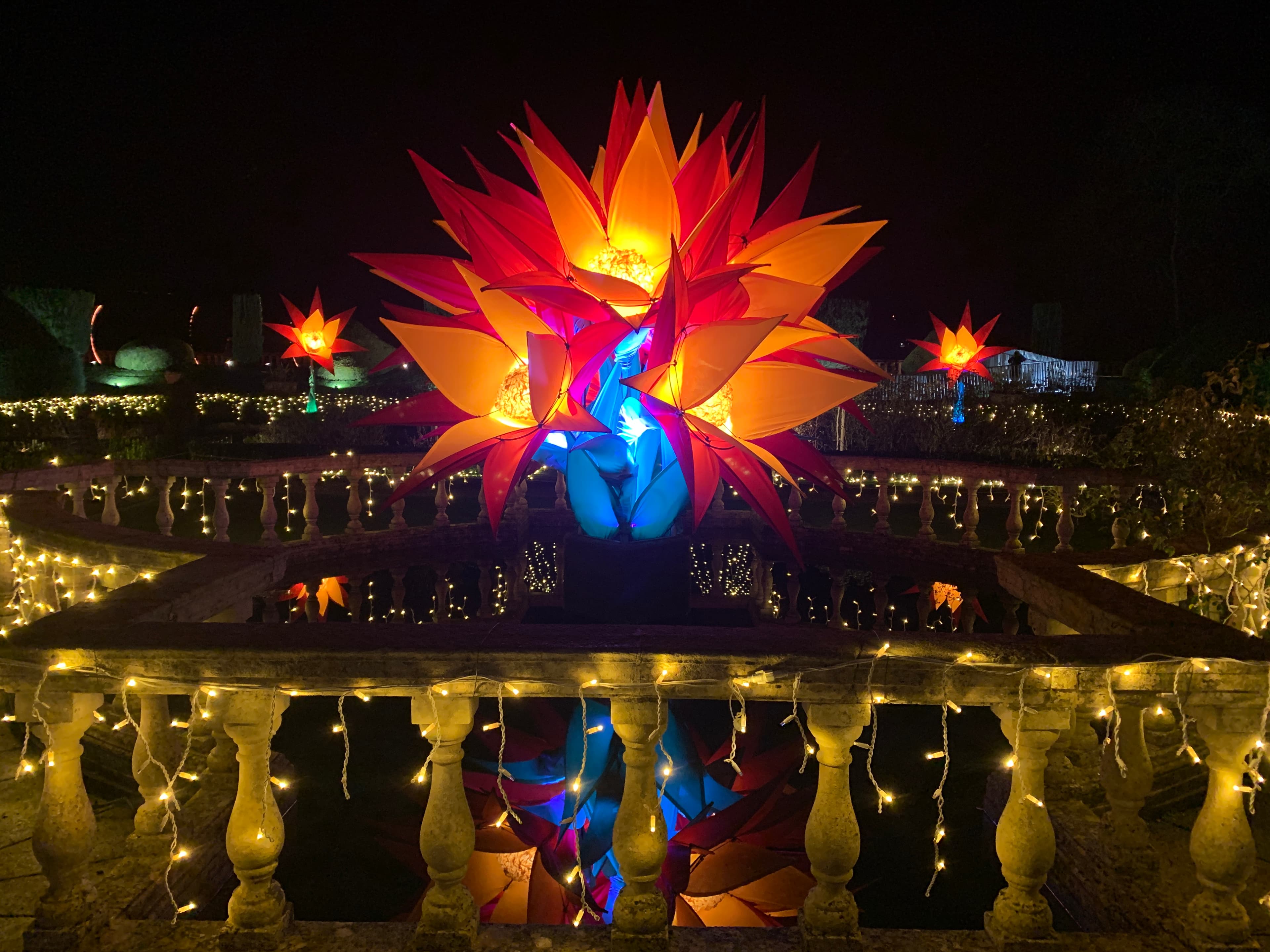 A vivid orange-and-red flower-shaped light sculpture reflected in a pool on Sudeley's terrace, fairy lights threaded through the stone balustrade