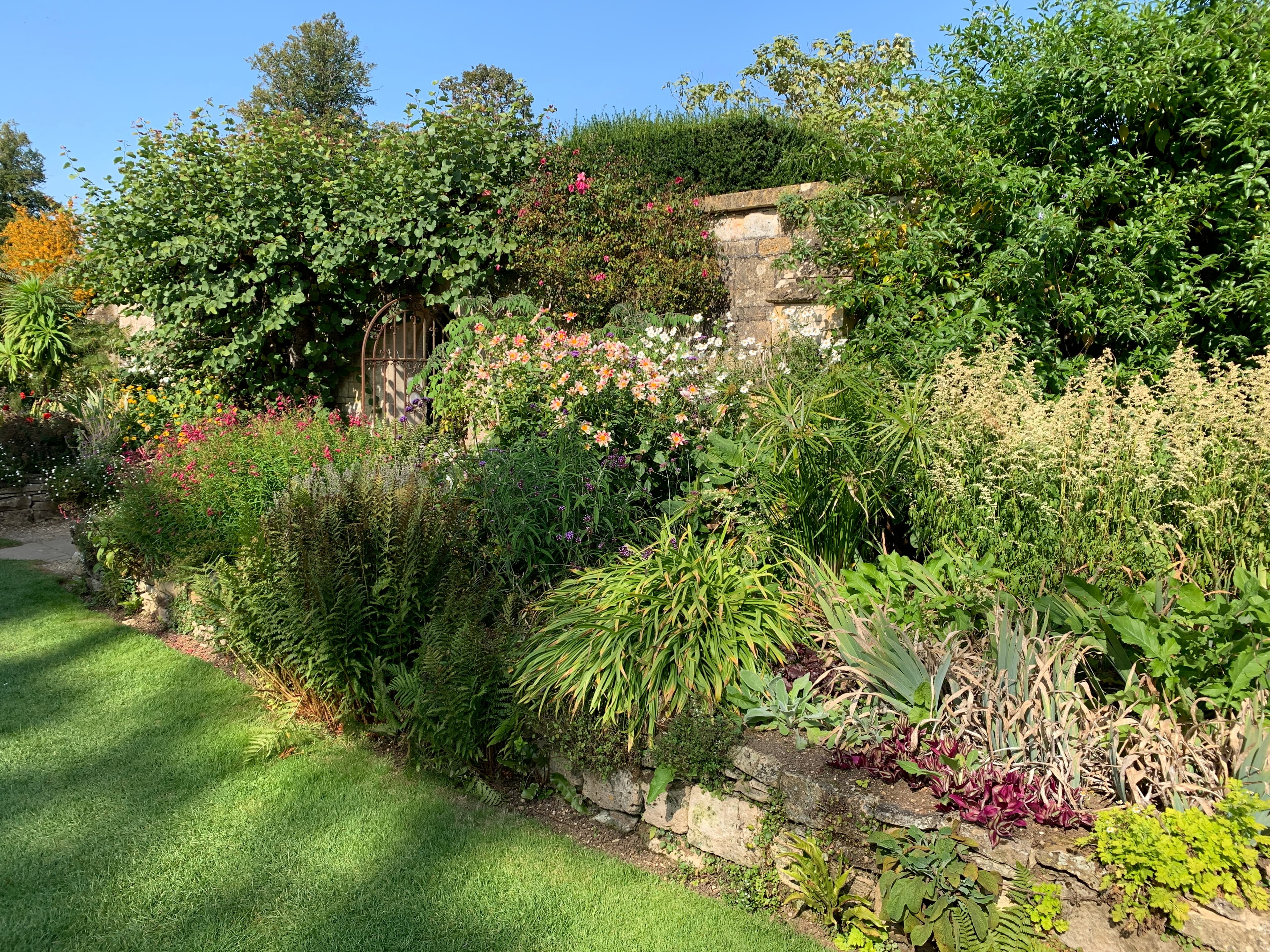 A late-summer herbaceous border at Sudeley — penstemons, daylilies and asters spilling over a Cotswold-stone wall under climbing roses