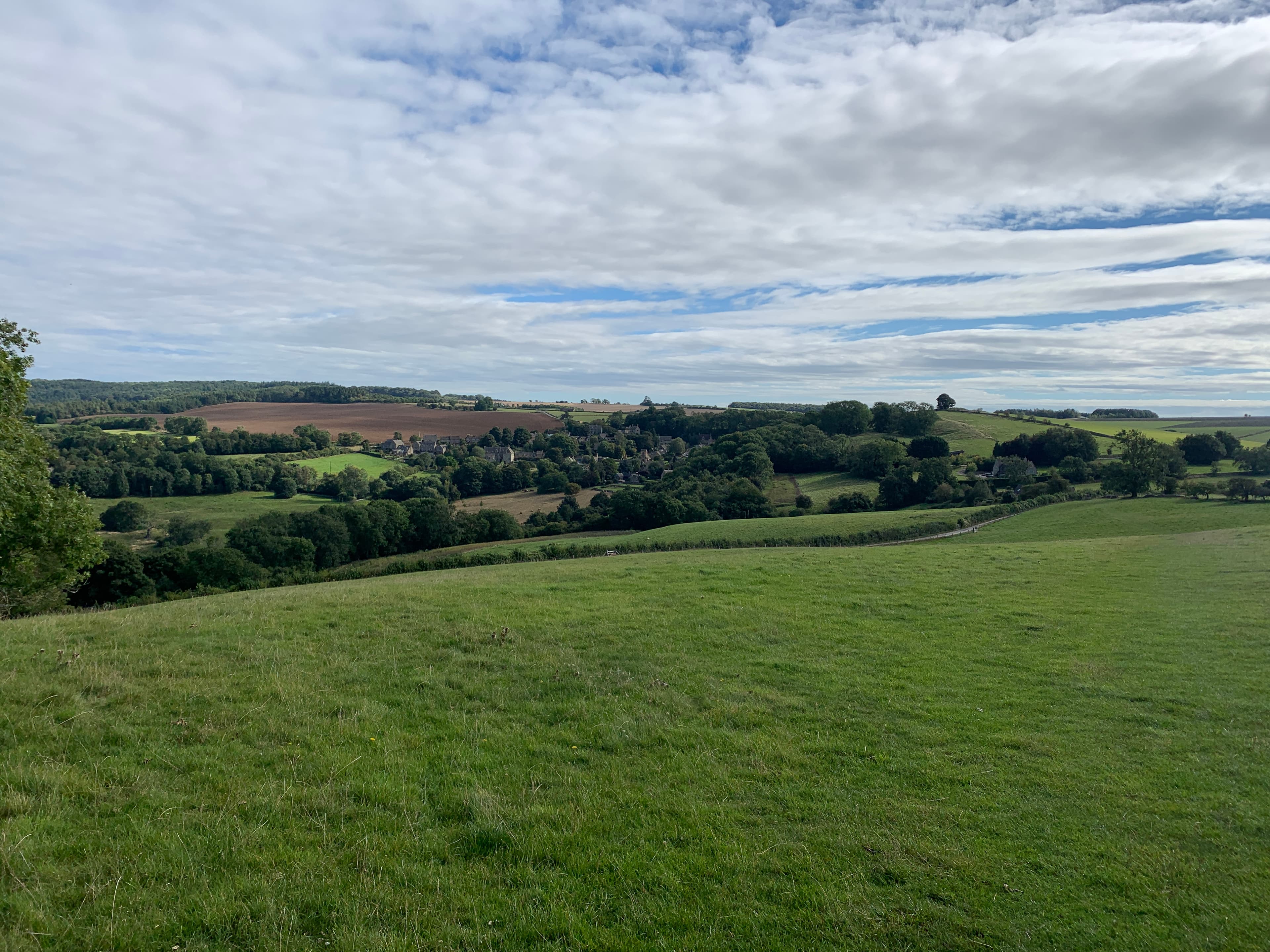 A wide Cotswold valley with a village nestled below, viewed from a hillside path