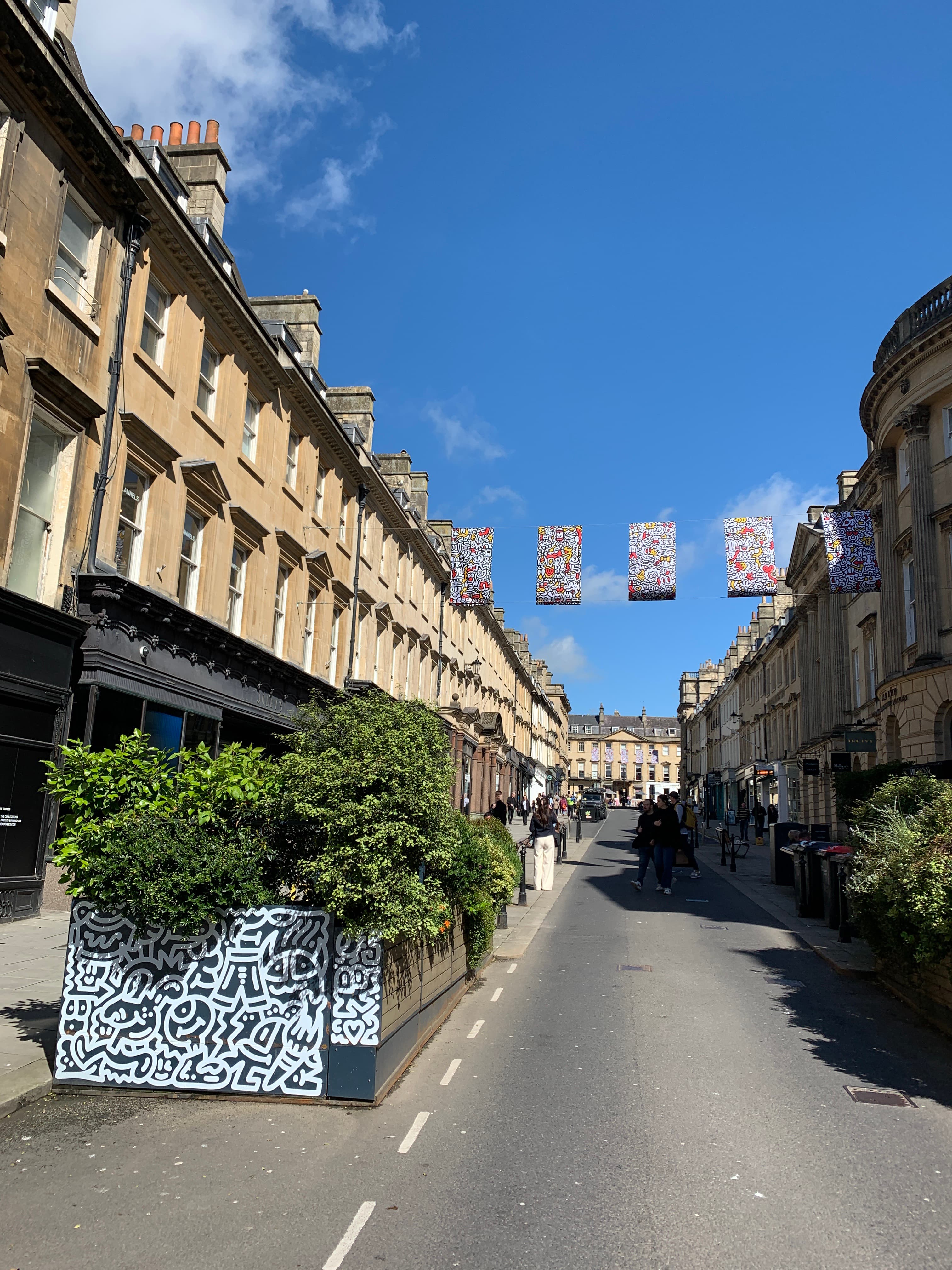A Bath side-street with colourful bunting strung between Georgian terraces and a large black-and-white mural on a corner building