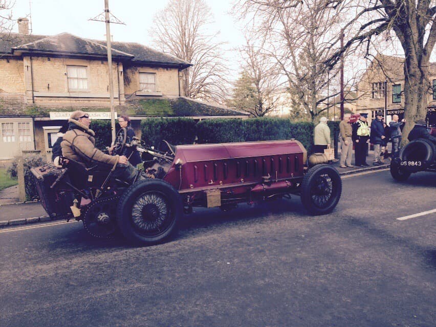 A vintage racing car driving through Bourton on a winter morning, with onlookers and another classic car following behind