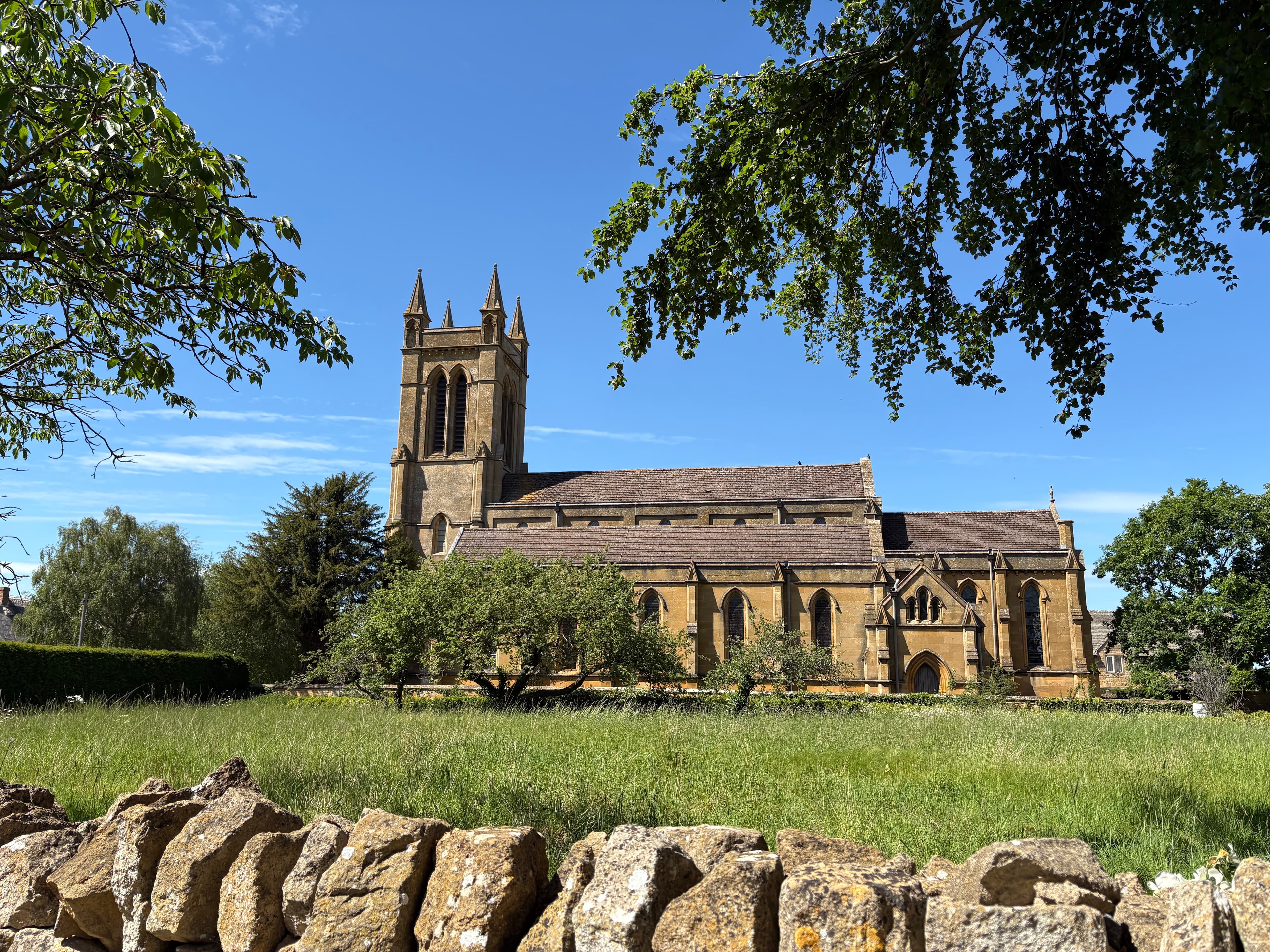 St Michael & All Angels, Broadway — sandstone church and tower seen across a wildflower meadow over a drystone wall