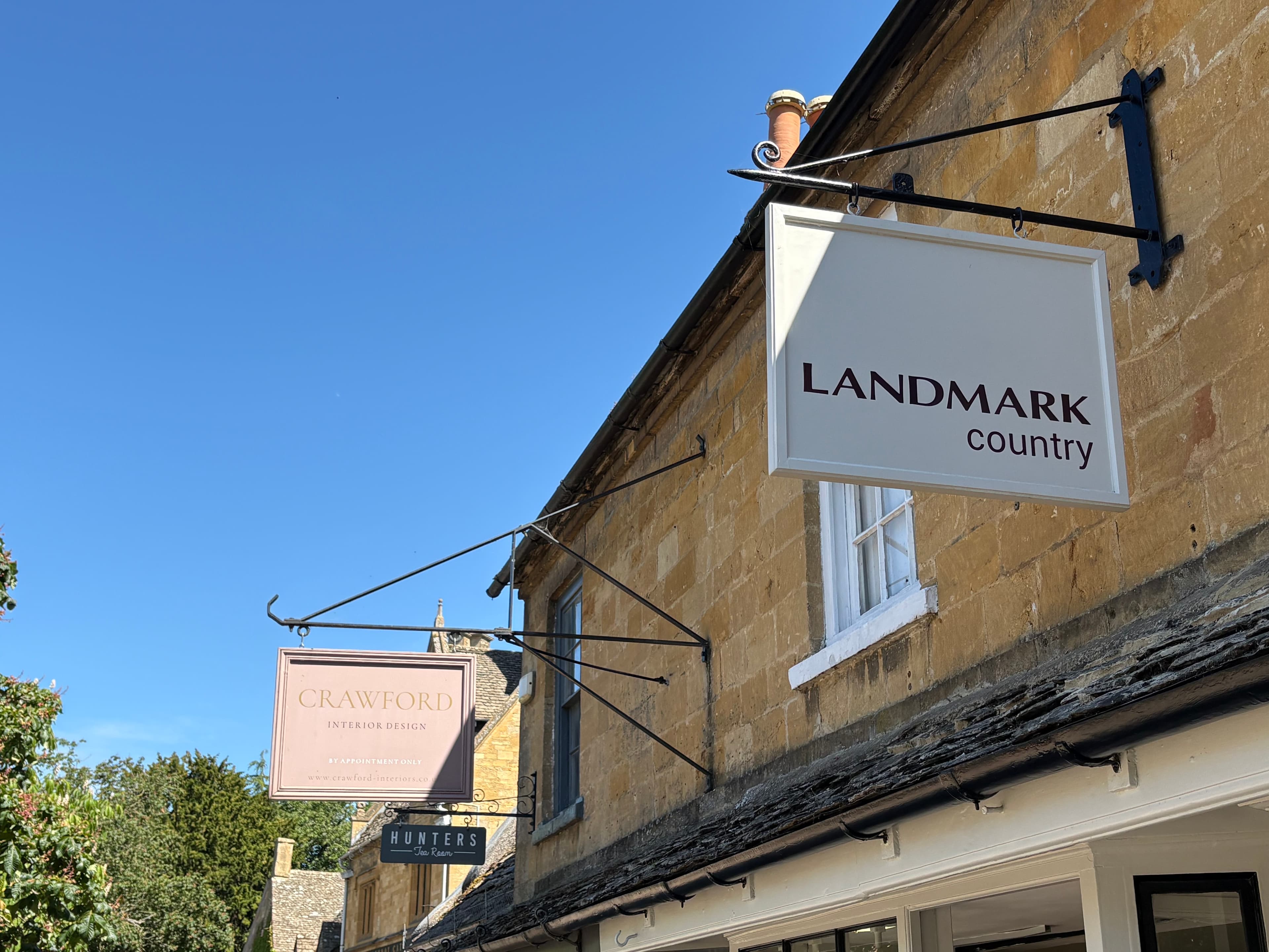 Hanging shop signs for 'Landmark country' and 'Crawford Interior Design' on a Cotswold-stone shopfront