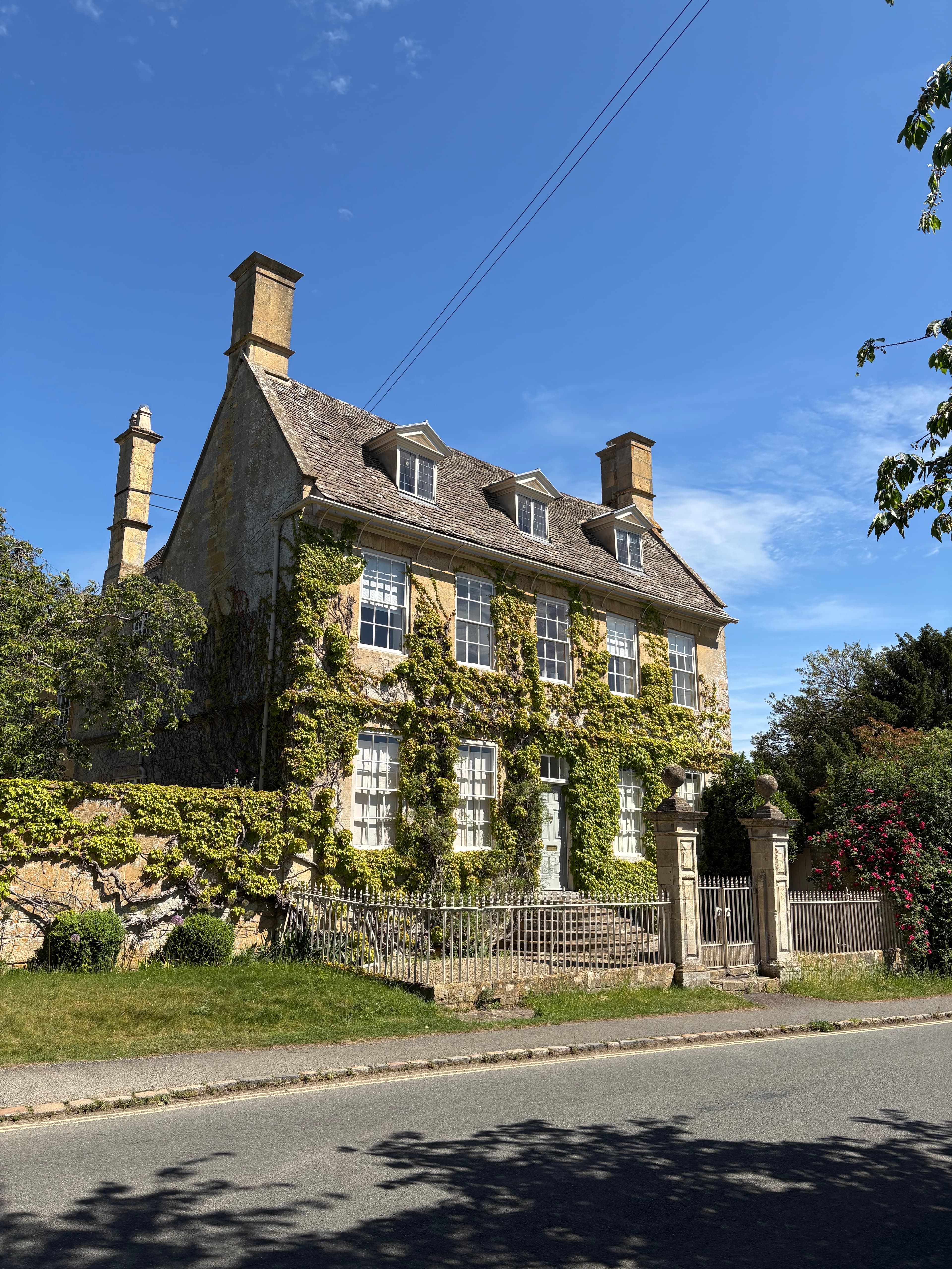 A long Cotswold-stone country house in Broadway with chimneys and dormer windows, climbing roses against the wall