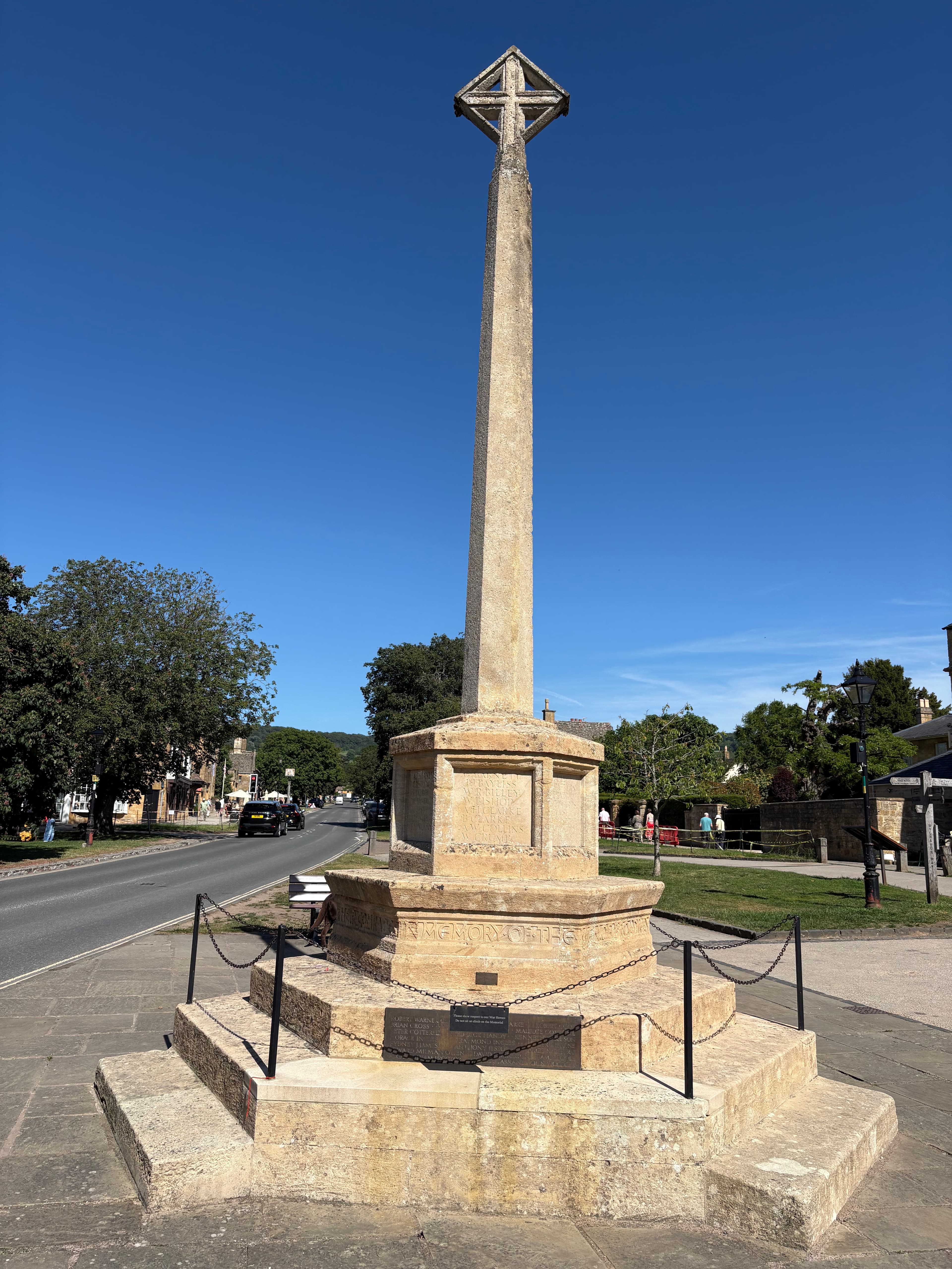The tall stone village cross on Broadway's high street, with the war memorial plaque at its base