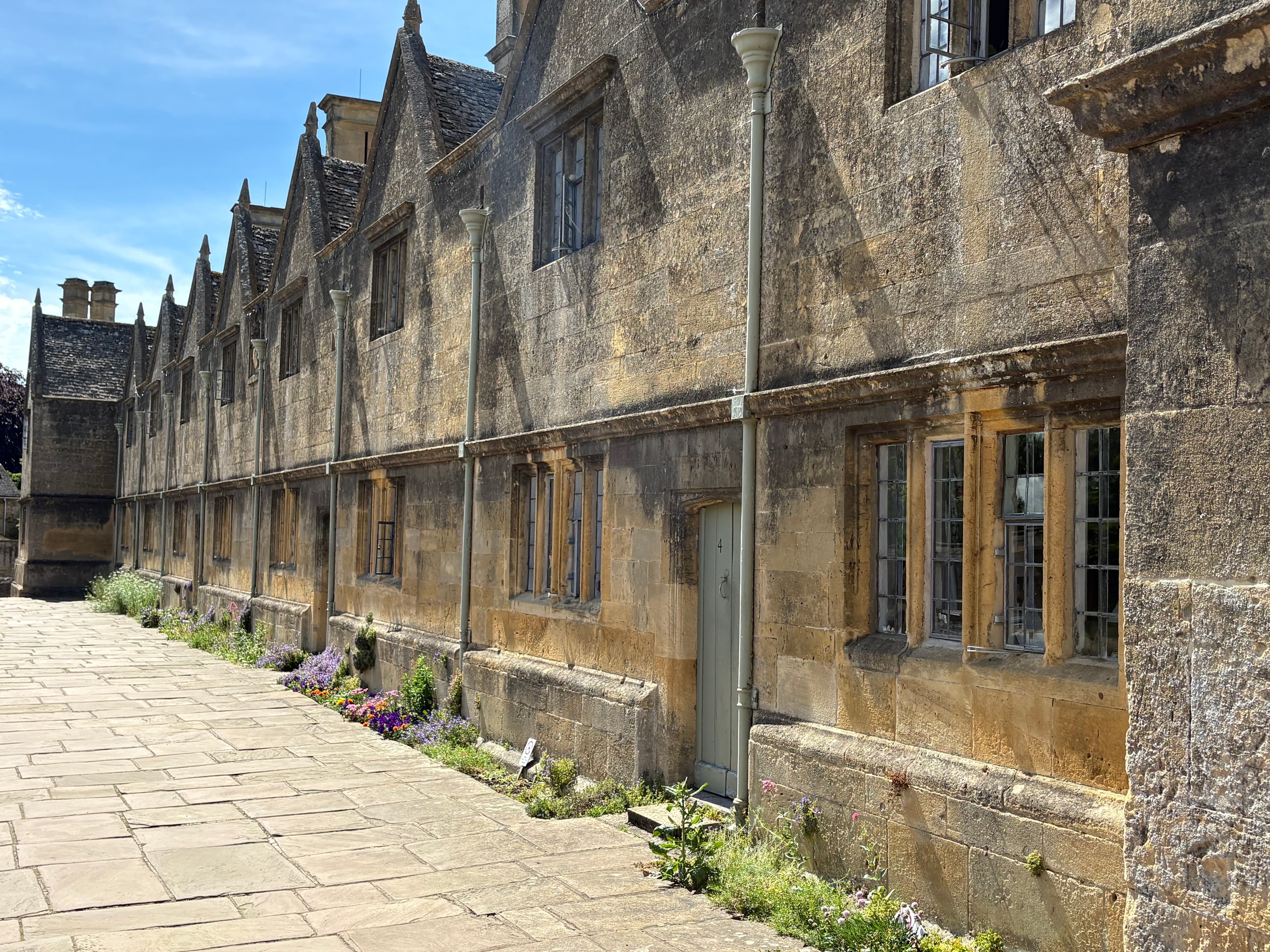 The 17th-century almshouses below the church — a long terrace of gabled stone cottages set above a pavement of wildflowers