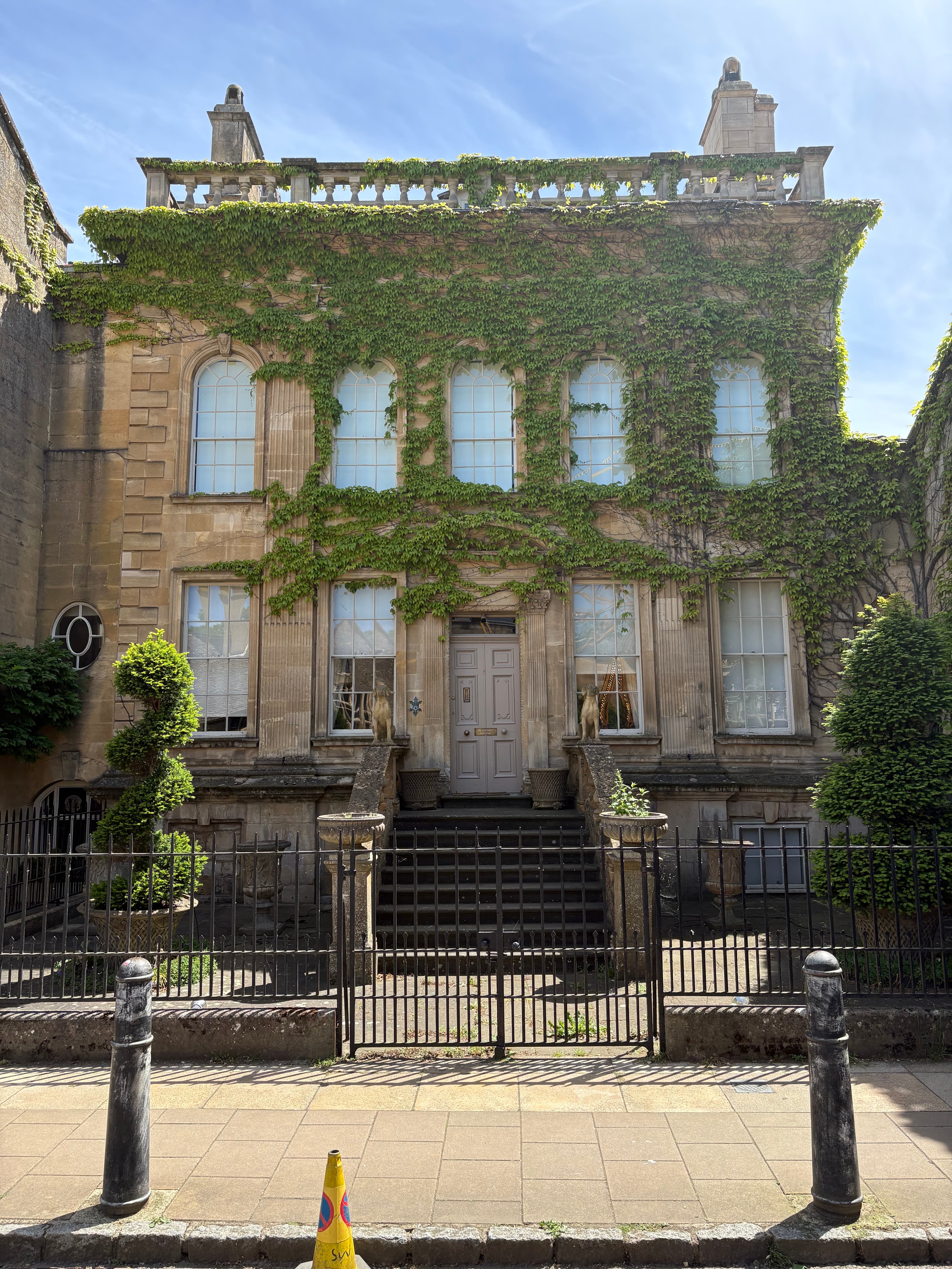 A handsome Cotswold-stone building thickly clad in ivy, with stone-mullion windows and clipped front hedges