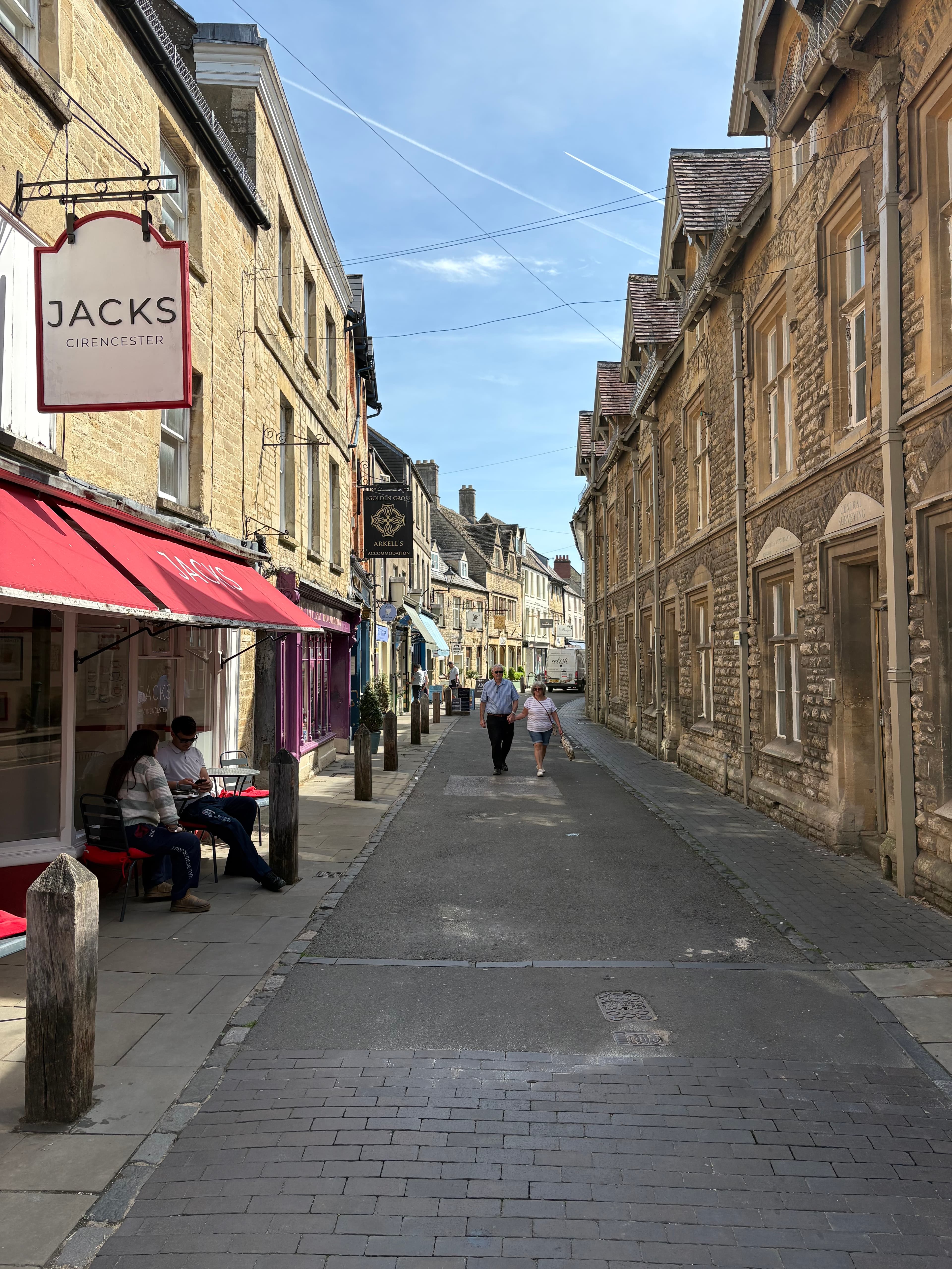 Looking up the high street under a long banner for Jacks Cirencester, with cafes spilling onto the pavement
